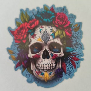 Peut inclure: Un crâne de sucre coloré avec une couronne de fleurs, comprenant des fleurs rouges, bleues et jaunes. Le crâne a des yeux noirs et un visage blanc avec des détails complexes.