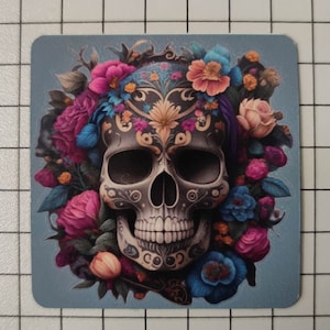 Peut inclure: Un crâne de sucre noir et blanc avec des motifs floraux complexes, entouré de fleurs colorées dans des tons de rose, bleu et jaune. Le crâne est décoré d'un motif floral et a un visage détaillé.