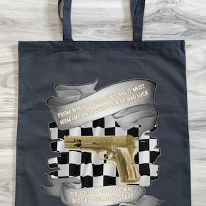 Op de afbeelding: Een zwarte tote bag met een geruit patroon en een gouden pistool. De tas heeft een wit lint met de tekst "From where you're kneeling it must seem like an 18-carat run of bad luck. The truth is, the game was rigged from the start."