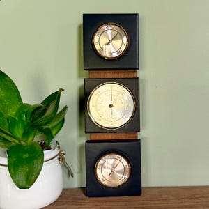 May include: A vertical weather station with three circular gauges. Each gauge has a gold-colored bezel and a white face with black markings. The gauges are set in black square frames and mounted on a wooden backing. A thermometer, barometer, and hygrometer are visible.
