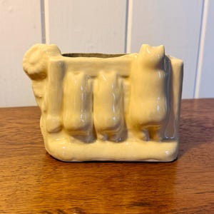 May include: A rectangular, light yellow ceramic planter with a whimsical design. The planter features three stylized cat figures and a ram's head on the sides. The planter is sitting on a wooden surface.