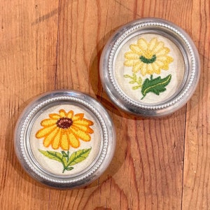 May include: Two silver framed coasters with embroidered yellow sunflowers on a white background. The sunflowers have green leaves and stems.