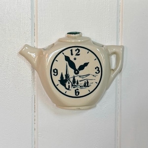 May include: Cream-colored teapot-shaped wall clock with a green accent on top. The clock face features black numerals and hands, with a scenic illustration of a church and mountains. The clock is mounted on a white wooden surface.