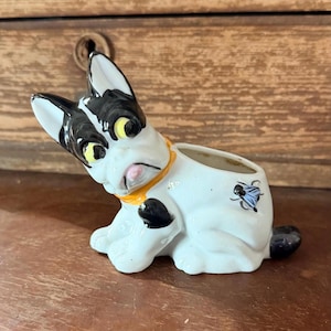 May include: A ceramic dog figurine in a sitting position, featuring a white body with black spots and accents. The dog has black ears, yellow eyes, and a pink tongue. It wears an orange collar and has a small opening on its back.