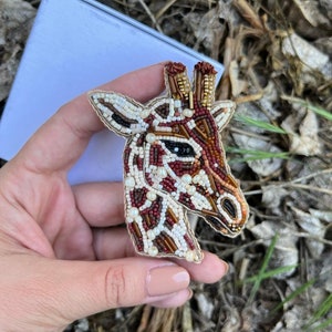 Peut inclure: Une broche en forme de girafe en perles de couleur marron, blanche et rouge. La t&ecirc;te de la girafe est tourn&eacute;e vers la droite. La broche est tenue dans une main.