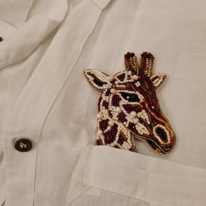 Peut inclure: Une broche en forme de girafe en perles de couleur marron, blanc et or, &eacute;pingl&eacute;e &agrave; la poche d'une chemise blanche.
