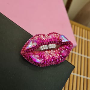 May include: A pink and iridescent beaded brooch shaped like a pair of lips. The lips are closed and have a row of white beads for teeth.