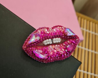May include: A pink and iridescent beaded brooch shaped like a pair of lips. The lips are closed and have a row of white beads for teeth.