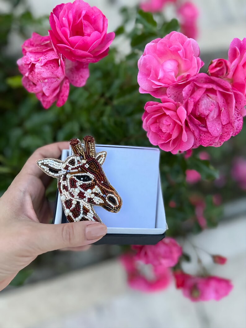 Peut inclure: Une broche en forme de girafe en perles de couleur marron, blanche et dor&eacute;e, pr&eacute;sent&eacute;e dans une bo&icirc;te cadeau noire et blanche, avec des roses roses en arri&egrave;re-plan.