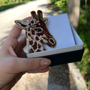 Peut inclure: Une broche en forme de girafe en perles de couleur marron, blanc et or. La broche est pr&eacute;sent&eacute;e dans une bo&icirc;te noire et blanche.