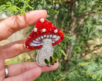 Peut inclure: Broche champignon faite à la main. Le chapeau rouge est orné de perles et de perles, le pied est blanc et argenté, et de petites feuilles vertes sont attachées. La broche est posée sur un fond de feuillage vert.