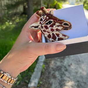 Peut inclure: Une broche en forme de girafe en perles de couleur marron, rouge et blanc. La broche est dans une petite bo&icirc;te avec un int&eacute;rieur blanc et un ext&eacute;rieur noir.