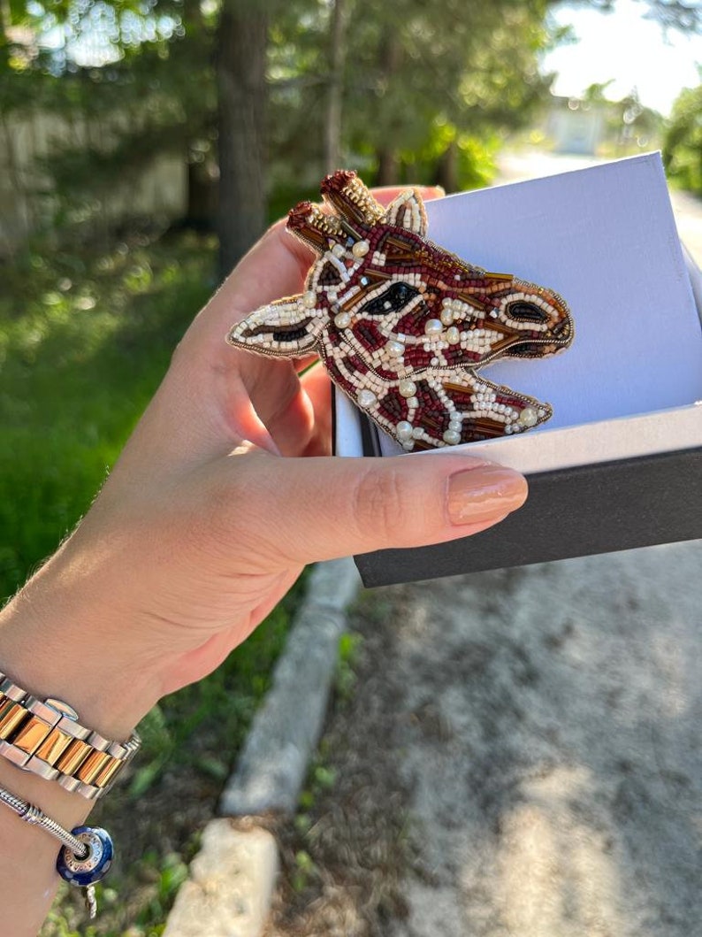 Peut inclure: Une broche en forme de girafe en perles de couleur marron, rouge et blanc. La broche est dans une petite bo&icirc;te avec un int&eacute;rieur blanc et un ext&eacute;rieur noir.