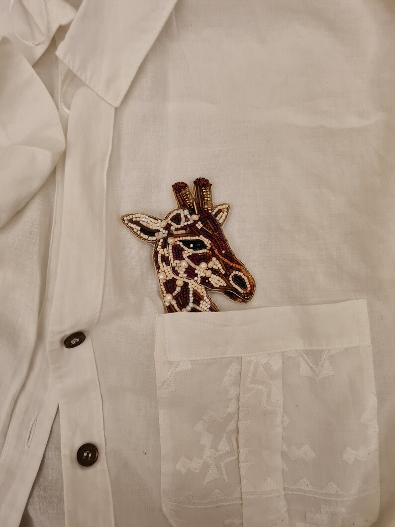 Peut inclure: Une chemise boutonn&eacute;e blanche avec une broche en forme de girafe en perles brunes et blanches &eacute;pingl&eacute;e &agrave; la poche.