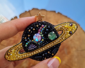 Peut inclure: Une broche en forme de planète avec une surface perlée noire, une étoile dorée au sommet et un anneau orné de cristaux jaunes. Le centre comprend un grand cristal irisé et un cristal vert plus petit, entourés de perles complexes.