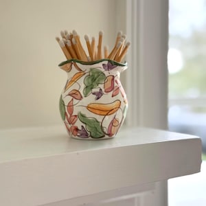 May include: A decorative ceramic match holder with a floral design in orange, green, and purple. The vase is filled with wooden matches. The match holder sits on a white shelf.