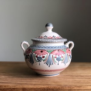 May include: A white ceramic sugar bowl with a lid, featuring a hand-painted floral design in shades of pink, blue, and green. The bowl has two handles and a rope-like design around the lid.