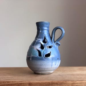 May include: A small, blue ceramic vase with a handle and a cut-out design of leaves. The vase has a white glaze on the bottom and a light blue glaze on the top.