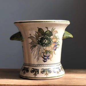 May include: A white ceramic vase with green handles and a floral design featuring fruits and vegetables. The vase has a decorative border around the top and bottom.