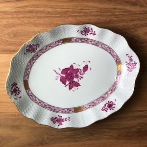 May include: An oval-shaped serving dish with a white background and a pink floral design. The dish has a gold rim and a scalloped edge.