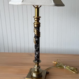 May include: A brass table lamp with a square base and claw feet. The lamp has a black and white patterned column with a gold spiral accent. The lampshade is a light beige color and is square-shaped.