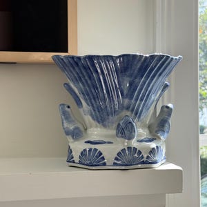 May include: A white ceramic planter featuring a blue hand-painted shell pattern. The vessel has a flared, scalloped rim and is decorated with three-dimensional sculpted figures of small seals positioned around the base, alternating with shell motifs.
