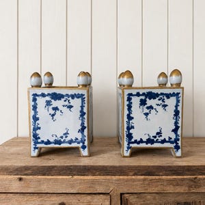 May include: A pair of square ceramic planters featuring a white base with blue floral vine patterns enclosed in a rectangular border. Each planter has four short feet and four decorative finials on the top corners, all finished with gold accents.
