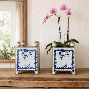 May include: Two square ceramic planters featuring a white base with blue floral patterns and gold-painted accents on the four top finials and base edges. One planter is empty, while the other contains a potted orchid with pink flowers and green leaves.