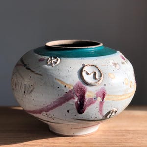 May include: A white ceramic vase with a teal blue rim and a variety of painted designs, including a swirl, circles, and dots. The vase has a rounded body and a slightly flared base.