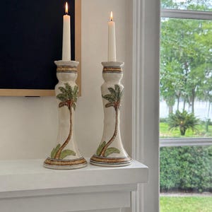 May include: Two cream-colored ceramic candle holders with palm tree designs. Each holder has a lit, slender white candle. The palm trees are depicted with green fronds and brown trunks. The holders sit on a white shelf.