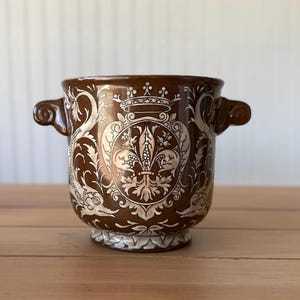 May include: A brown ceramic planter with two side handles, featuring a white embossed fleur-de-lis emblem topped with a crown. The exterior is decorated with intricate white scrollwork and stylized dolphin motifs encircling the central crest.