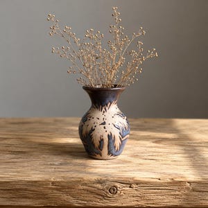 May include: A small, handcrafted ceramic vase with a dark blue rim and a beige body decorated with a blue leaf pattern. The vase holds dried, light brown stems and sits on a rustic wooden surface.