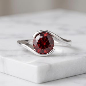Può includere: Un anello color argento con una pietra preziosa rotonda e sfaccettata di colore rosso intenso, fissata con una montatura a bypass. La fascia si curva armoniosamente attorno alla pietra centrale, creando una cornice metallica lucida.
