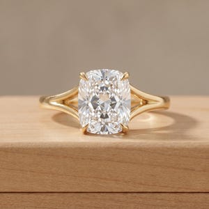 Puede incluir: Un anillo de oro con una piedra grande, transparente y de corte cojín, sujeta por cuatro garras. La banda tiene un diseño de brazo dividido que se estrecha hacia el engaste, con un acabado metálico pulido que refleja la luz en su superficie.