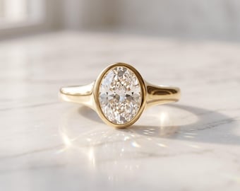 May include: A gold ring featuring a central oval-cut clear gemstone set in a smooth bezel setting. The band is polished yellow gold with a tapered design that connects to the oval setting, creating a clean and continuous metallic appearance.