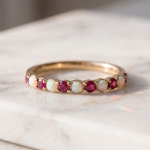 Puede incluir: Un anillo de banda de oro con un patrón alterno de ópalos blancos iridiscentes redondos y rubíes de color rojo intenso engastados en garras compartidas. Las piedras están dispuestas en una fila continua a lo largo de la parte frontal del anillo.