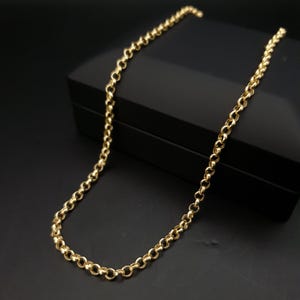 Puede incluir: Un collar de cadena de color dorado con un diseño de eslabones circulares. El collar se exhibe sobre una superficie negra, creando un contraste que resalta el brillo de la joya. La cadena es de material metálico brillante.