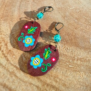 Op de afbeelding: Een paar houten oorbellen met een bloemenmotief. De oorbellen zijn geschilderd met blauwe, groene en rode bloemen op een bruine achtergrond. Elke oorbel heeft een turquoise kraal die eraan hangt.