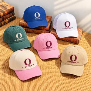 May include: Six baseball caps in various colors including blue, white, green, pink, beige, and a two-tone cream and pink design. Each cap features a large letter O embroidered above the name OLIVIA in a centered, horizontal layout.