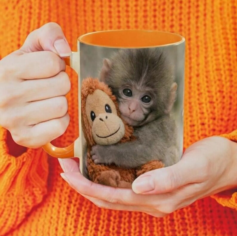 Puede incluir: Taza de cer&aacute;mica con un beb&eacute; mono y un mono de peluche. La taza tiene un interior y un asa de color naranja. La imagen la sostiene una persona que lleva un jersey naranja.
