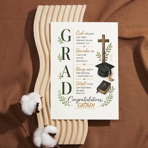 May include: A graduation card with the word "GRAD" in large letters, surrounded by floral accents. The card includes religious text and illustrations of a cross, graduation cap, and book. The card is displayed on a wooden stand with cotton accents.
