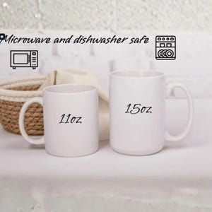 Puede incluir: Dos tazas de cer&aacute;mica blancas, una con la etiqueta "11oz" y la otra "15oz". Las tazas tienen un dise&ntilde;o sencillo con un asa curvada. El texto encima de las tazas dice "Microwave and dishwasher safe" con iconos relacionados.