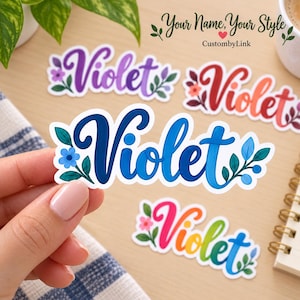 May include: A die-cut vinyl sticker featuring the name Violet in a bold blue script font, accented with small blue flowers and green leaves. The sticker is held up, with other similar stickers in purple, red, and rainbow gradients visible.
