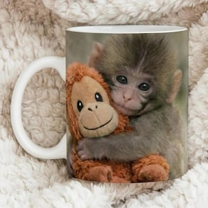 Puede incluir: Taza de cer&aacute;mica blanca con un mono beb&eacute; abrazando a un peluche de orangut&aacute;n. El mono tiene pelaje gris y ojos oscuros, mientras que el juguete es naranja con una cara sonriente. La taza tiene un asa blanca.
