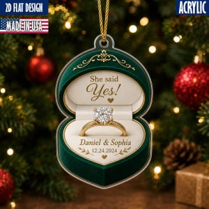 May include: A flat acrylic Christmas ornament shaped like an open dark green ring box with gold trim. Inside, a gold engagement ring with a round diamond sits on a cream cushion. Text reads: She said Yes!, Daniel & Sophia, and 12.24.2024.
