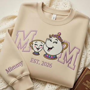 May include: Beige sweatshirt with embroidered design. Features the word "MOM" in purple, with a teacup and teapot illustration. The text "EST. 2026" is below the illustration. The sleeve cuff has the word "Mimmy" embroidered in purple.