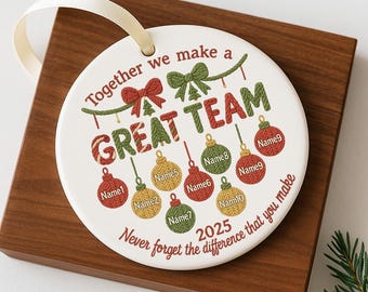 May include: A white ceramic ornament with the text "Together we make a GREAT TEAM" and the year 2025. The ornament features red, green, and gold-colored Christmas ornaments with names. It has a cream-colored ribbon for hanging.