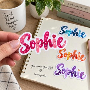 May include: A collection of die-cut vinyl stickers featuring the name Sophie in a cursive script font. Each sticker displays a unique color gradient filled with small heart patterns, including pink, blue, orange, and purple variations.