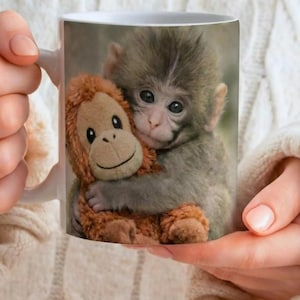 Puede incluir: Taza de cer&aacute;mica blanca con un beb&eacute; mono abrazando un peluche de orangut&aacute;n. El mono tiene pelaje gris y ojos grandes y oscuros. El peluche de orangut&aacute;n es marr&oacute;n y naranja. La taza la sostiene una persona que lleva un jersey blanco.