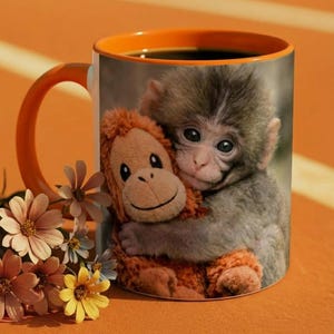 May include: A ceramic coffee mug with an orange handle and interior, filled with dark liquid. The mug features a print of a monkey hugging a stuffed animal. A small bouquet of flowers sits near the mug.